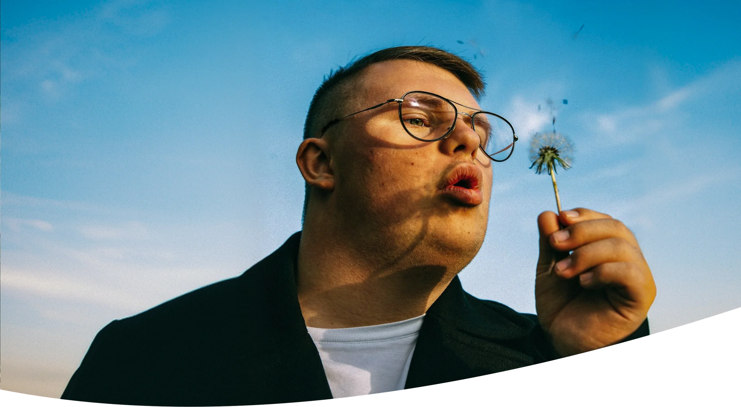 A man blows a dandelion against a blue sky, the home page hero photograph, cut with the brand's wave
